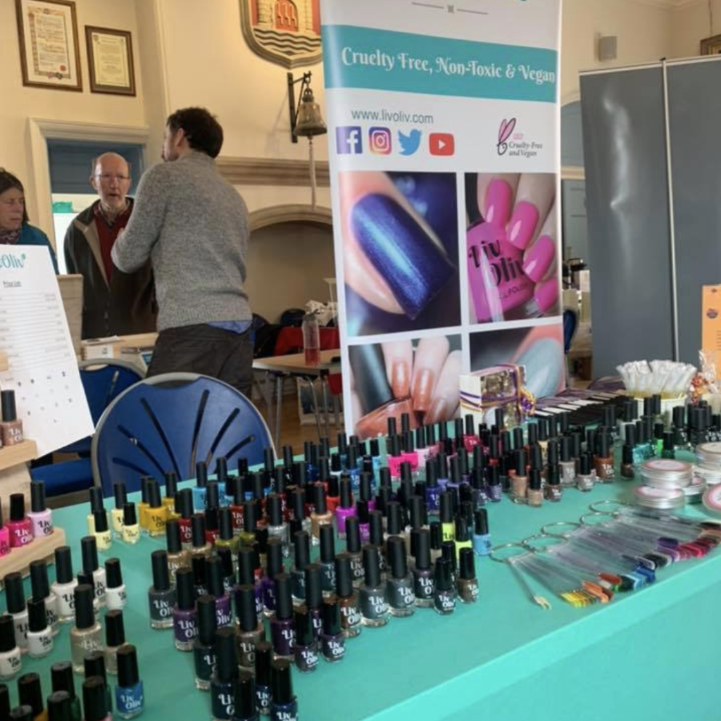 LivOliv booth at a community event, featuring rows of colorful nail polish bottles and a banner promoting cruelty-free, non-toxic, vegan products